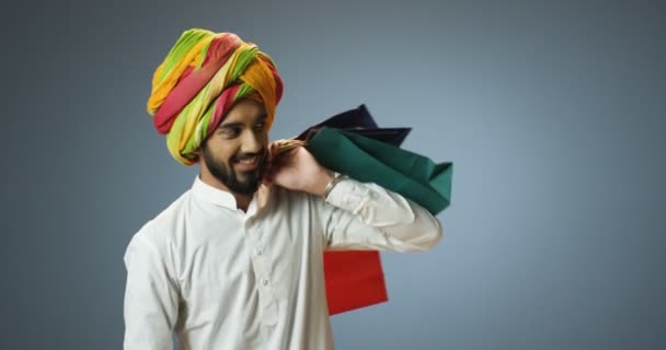 Όμορφος χαρούμενος νεαρός Ινδός με παραδοσιακό τουρμπάνι κρατώντας πακέτα πάνω από τον ώμο του μετά τα ψώνια και χαμογελώντας. Ευτυχισμένος Ινδός με χαμόγελο και τσάντες στο χέρι. - Πλάνα, βίντεο