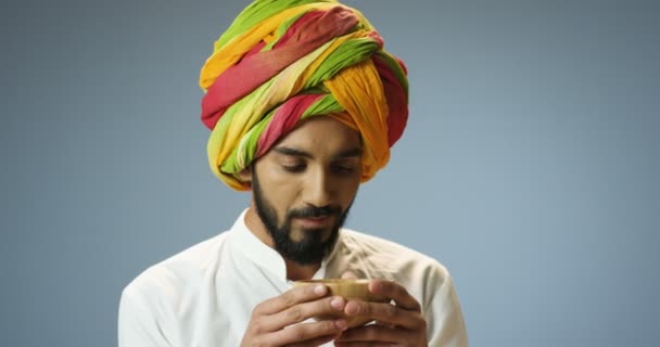 Primer plano de joven hindú guapo en turbante colorido tradicional y con barba sorbiendo bebida caliente de la taza. Atractivo macho indio bebiendo té sobre fondo gris de la pared. - Imágenes, Vídeo