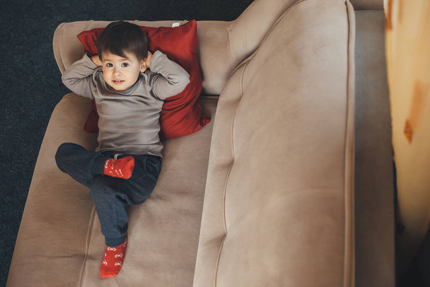 Vista superior de la foto de un niño caucásico feliz sonriendo a la cámara mientras yacía en la cama aburrido de aislamiento con calcetines rojos y usando una almohada - Foto, Imagen