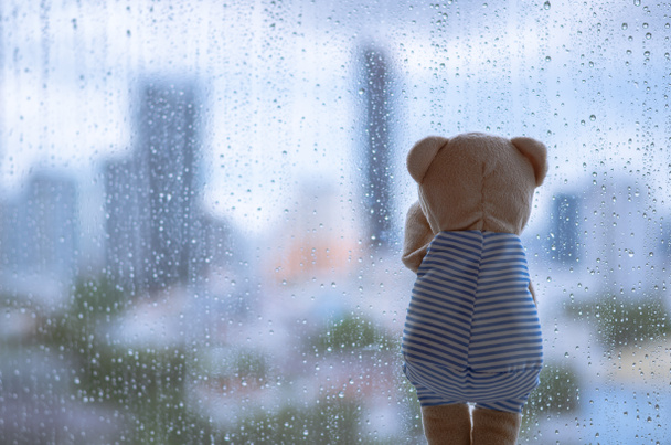 Teddy Bear płacze samotnie w oknie, gdy pada deszcz z zamazanym tłem miasta. - Zdjęcie, obraz