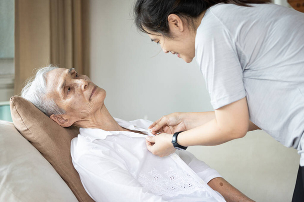 Asiatische Pflegerin, die älteren Patienten beim Anziehen hilft, Knopf am Hemd oder Kleiderwechsel für eine gelähmte Person, ältere Frau mit Lähmungen der Gliedmaßen, des Körpers oder der Muskeln Schwäche - Foto, Bild