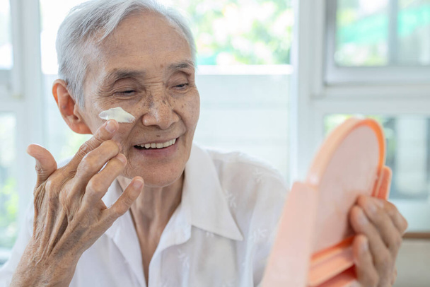 Glücklich lächelnde asiatische Seniorin cremt sich mit Sonnencreme auf die Wange ein, schützt ihr schönes Gesicht, ältere Menschen mit Hautpflegeprodukt für empfindliche Haut, Sonnencreme auf Gesichtshaut bei sonnigem Tag - Foto, Bild