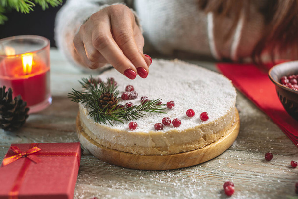 A woman is decorating a white mousse festive cake with red berries sprinkled with coconut flakes that imitating snow. Christmas and New year concept. - Photo, image