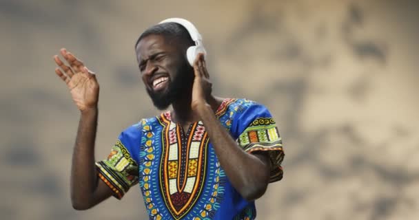 Jeune homme afro-américain beau avec barbe et en vêtements traditionnels chantant et écoutant de la musique dans des écouteurs. Guy appréciant la chanson préférée. - Séquence, vidéo
