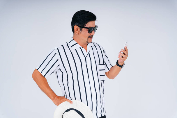 Портрет путешественника пожилого пожилого мужчины в белой рубашке воротничка носить солнцезащитные очки, держа шляпу и смартфон. - Фото, изображение