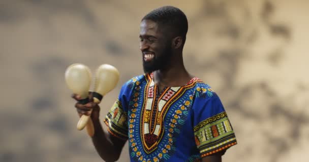 Egy jóképű afro-amerikai férfi portréja hagyományos ruhákban, vidáman mosolyog és maracán játszik. Boldog srác kezében hangszerek és így rímel a zene, miközben remeg. - Felvétel, videó
