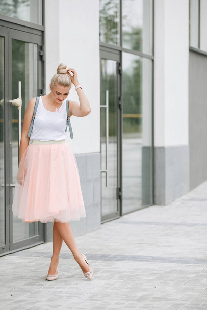 Blondynka pozująca na ulicy miasta. Modne ubrania, spójrz - Różowa spódnica, biała bluzka i szary plecak. - Zdjęcie, obraz