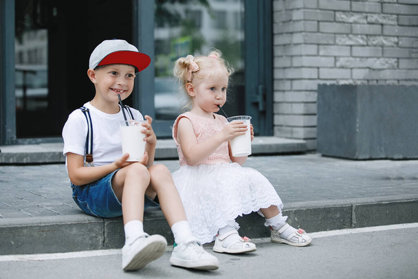 Blonde and the boy with a milkshake on the curb on a city street. A brother in a baseball cap and a sister with ponytails in pink sit on the sidewalk. - Foto, afbeelding