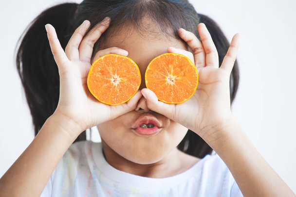 かわいいアジア系の子供の女の子は、彼女の目を覆う新鮮なオレンジを保持。新鮮な果物と遊ぶのが楽しい子供. - 写真・画像