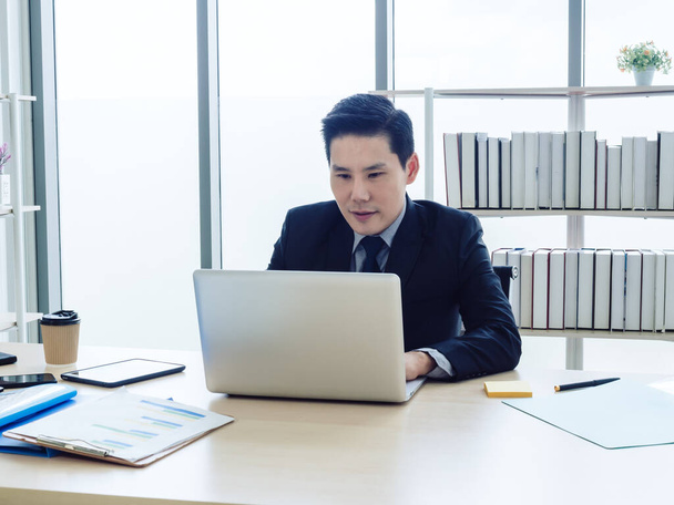 Asian businessman in suit sitting at table and working on laptop computer in office. Manager watching and typing on laptop computer in well-lit workplace. Business, technology and finance concept. - 写真・画像