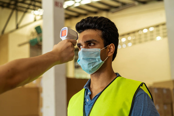 Young Indian factory warehouse worker wearing face mask getting body temperature check with infrared thermometer scanner before working. Security measure at workplace in Coronavirus Covid-19 pandemic - Foto, Imagen