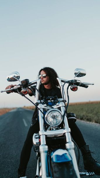 Невеликий фокус стильного кучерявого чоловіка з сонцезахисними окулярами, що позує на мотоциклі на дорозі
 - Фото, зображення