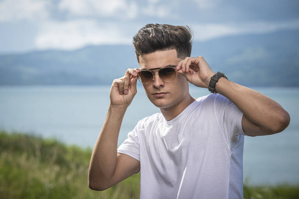 A closeup portrait of an attractive young male with a nature background behind putting his sunglasses on - Φωτογραφία, εικόνα