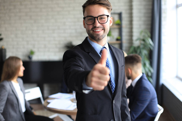 Happy businessman showing his thumb up with colleagues in the background in office - Foto, imagen