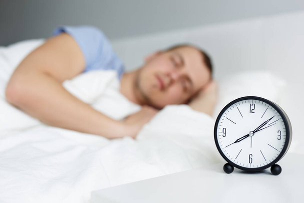 rentoutumista ja aikaa käsite - lähikuva herätyskello yöpöydällä ja nuori mies nukkuu sängyssä kotona - Valokuva, kuva