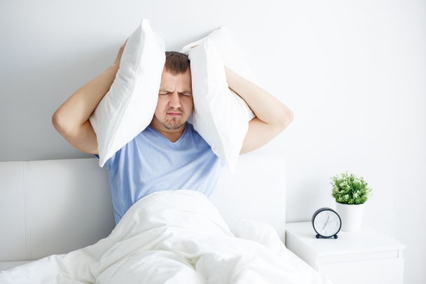 Derpression, Schlaflosigkeit und Kopfschmerz - Porträt eines müden jungen Mannes, der auf dem Bett sitzt und Kopfkissen hält - Foto, Bild