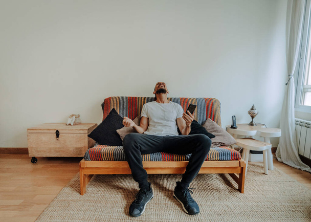 A Hispanic man sitting on the couch with a smartphone in his hands and relaxing - Foto, imagen