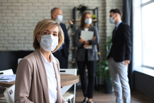 Businesswoman in medical mask with her staff, people group in background at modern bright office indoors - Fotoğraf, Görsel