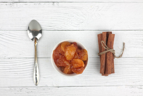bowl of apple jam, spoon and cinnamon sticks on white wooden background flat lay. Image contains copy space - Fotografie, Obrázek