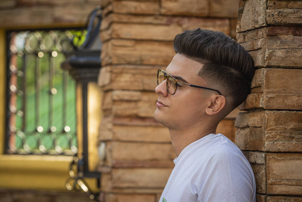 A closeup portrait of a handsome young man with a trendy hairstyle and sunglasses leaning on a stone wall - Fotoğraf, Görsel