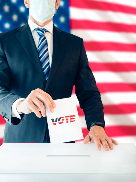 Hombre vestido con un traje y máscara protectora votando en las elecciones para presidente con la bandera de Estados Unidos detrás de él, enfoque selectivo - Foto, Imagen