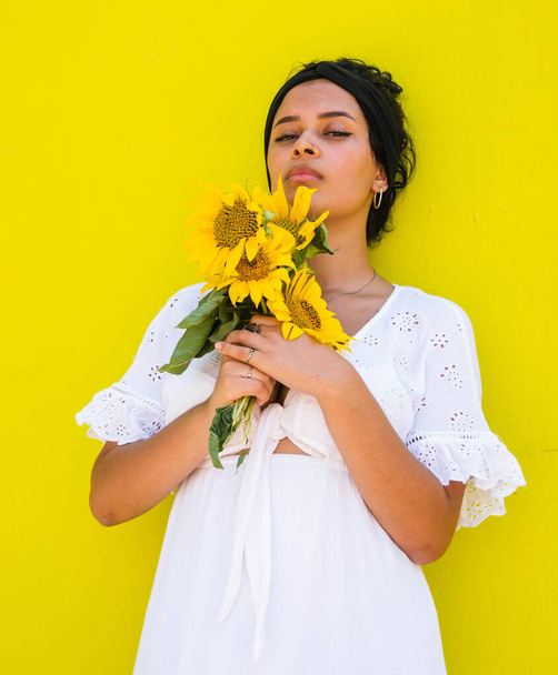 A vertical portrait of an attractive young female with sunflowers posing against a yellow background - Fotoğraf, Görsel