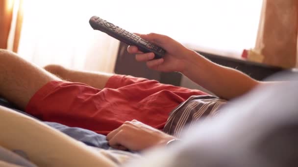Уставший человек лежит на кровати и эмоционально смотрит телевизор с дистанционным управлением - Кадры, видео