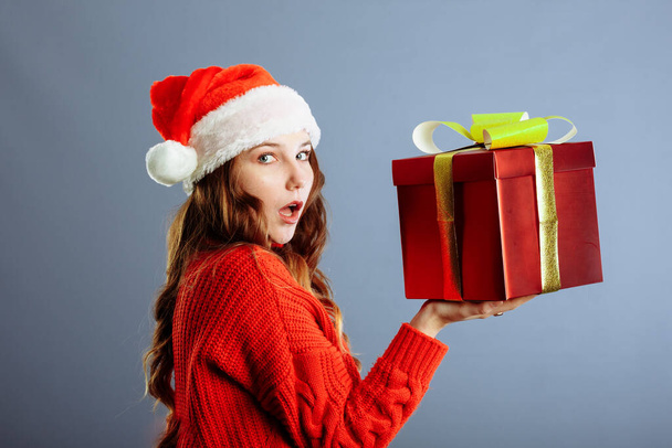 Beautiful Caucasian girl in dress and Santa hat is holding presents, looking at camera and smiling, isolated on gray background.. - Φωτογραφία, εικόνα