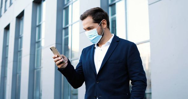 Caucasian young handsome man in medical mask tapping and scrolling on mobile phone outside at business center. Happy businessman in respiratory protection exting message on smartphone. Pandemic. - 写真・画像
