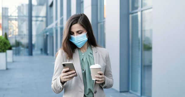 Young Caucasian pretty businesswoman in medical mask walking the street at business center with coffee to-go and texting message on smartphone. Beautiful woman tapping and scrolling on mobile phone. - Foto, Imagen