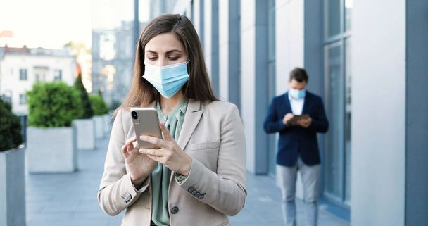 Young Caucasian pretty businesswoman in medical mask standing at street at business center and texting message on smartphone. Beautiful woman tapping and scrolling on mobile phone. Man on background. - Foto, Bild