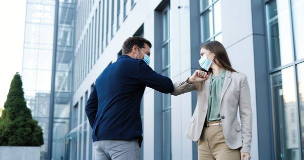 Caucasian young joyful female and male in medical masks meeting at street and greeting with elbows. Happy businessman and businesswoman talking and chatting cheerfully. Couple of friends. - Fotografie, Obrázek