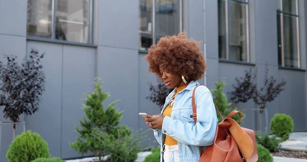 African American young stylish curly woman tapping or scrolling on smartphone and walking at city street. - Photo, image