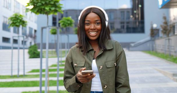 Cheerful young African American beautiful woman in headphones smiling cheerfully at city street and listening to music on smartphone. - Zdjęcie, obraz