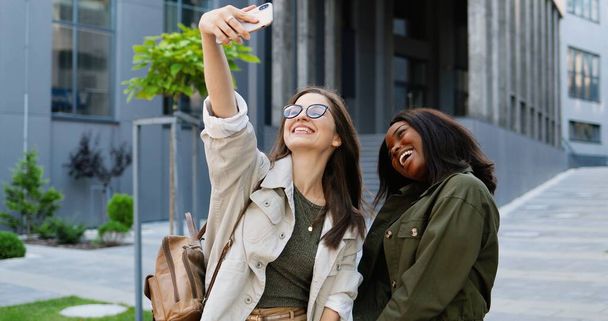Mixed-races young pretty females smiling cheerfully to camera and taking selfie photo with mobile phone camera at city street. - Fotó, kép