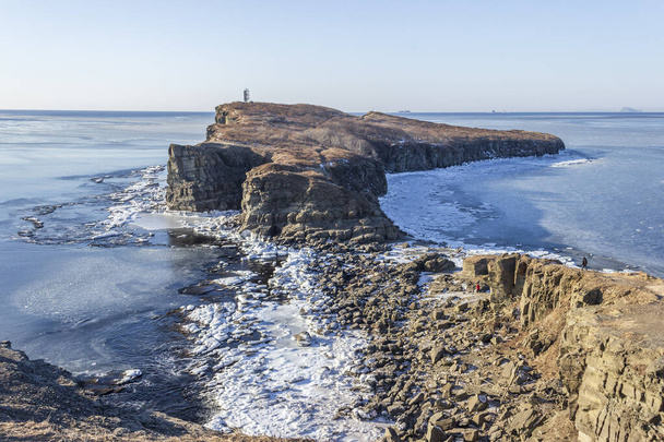 Skaliste wybrzeże Przylądka Tobizin na rosyjskiej wyspie we Władywostoku, pokryte śniegiem i pływającym lodem, w słoneczny zimowy dzień. - Zdjęcie, obraz
