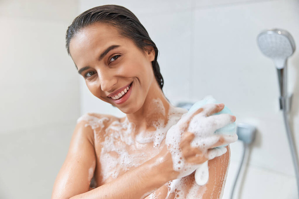 Urocza młoda kobieta używa złuszczającej gąbki podczas brania prysznica - Zdjęcie, obraz