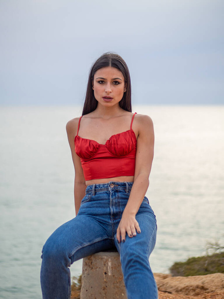 A beautiful young female wearing a sexy red crop top and jeans and sitting on a rock with the sea in the background - Fotó, kép