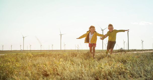 Κοριτσάκι και αγόρι τρέχουν μπροστά στους ανεμόμυλους. Ανανεώσιμες πηγές ενέργειας και αειφόροι πόροι - ανεμόμυλοι. παιδιά που παίζουν με τον άνεμο κοντά σε ανεμογεννήτρια. - Φωτογραφία, εικόνα