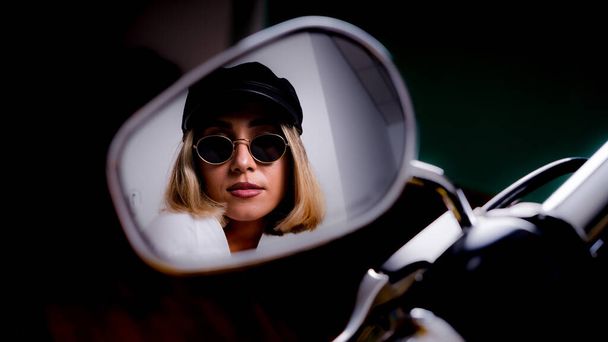 A cool blonde female with sunglasses and a hat looking in the mirror of a motorcycle - Zdjęcie, obraz