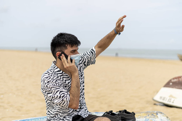 Güneşli bir günde plajda telefonla konuşurken koruyucu maske takan İspanyol bir adam. - Fotoğraf, Görsel