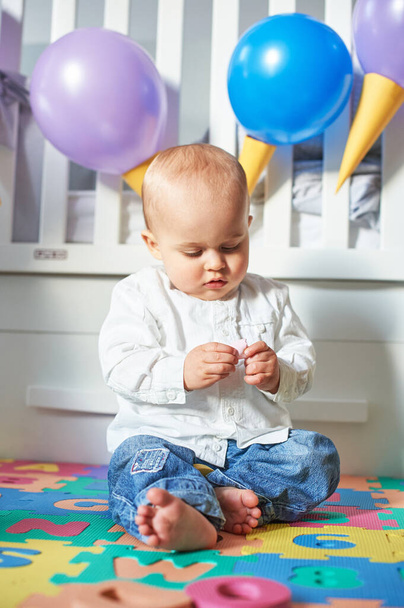 A baby sitting on the floor with a colorful carpet and with the crib decorated with balloons in the background and a candy in the hand - Photo, image