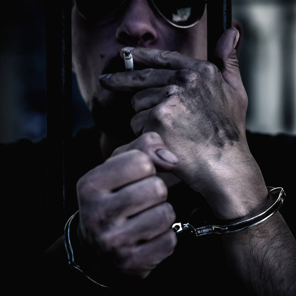 Close up portrait of arrested and handcuffed man imprisoned for crime smokes from behind bars. Gangster concept.  - Photo, Image