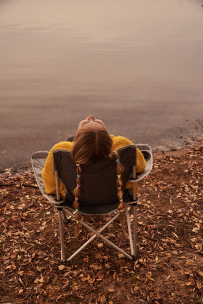 A teenage girl in a bright yellow sweater is sitting in a camp chair on the shore of a lake. - Φωτογραφία, εικόνα