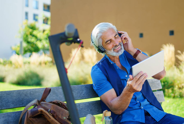 Portrait of mature man with headphones sitting outdoors in city, using tablet. - Фото, изображение