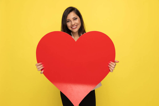 happy young girl in pink suit holding big red heart and smiling on yellow background - Zdjęcie, obraz