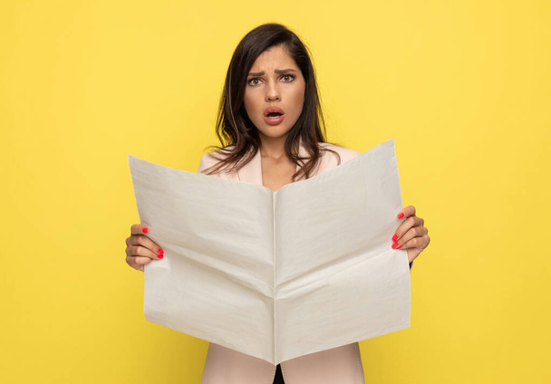 zaklatott fiatal nő rózsaszín öltöny nyitó száj sokkolva módon és olvasás újság, álló sárga háttér - Fotó, kép
