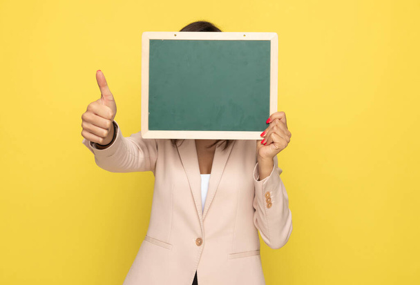 young businesswoman in pink suit covering face with blackboard and making thumbs up gesture, standing and posing on yellow background - Photo, image