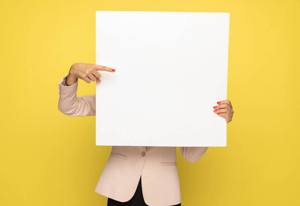 young fashion model in pink suit covering face with empty board and pointing finger to side on yellow background - Foto, Bild