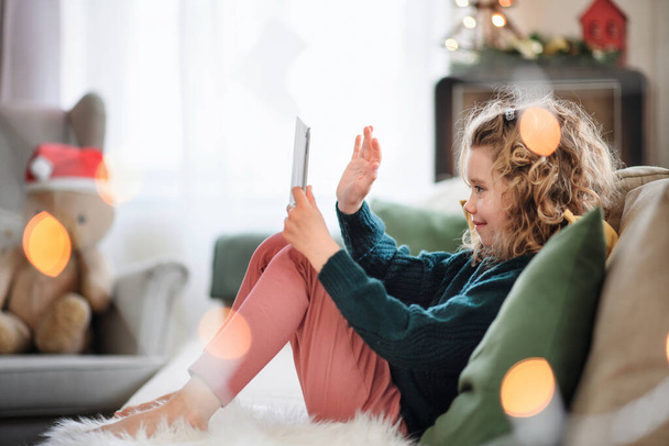 Kleines Mädchen mit Tablet sitzt zu Weihnachten zu Hause und hat Videoanruf. - Foto, Bild